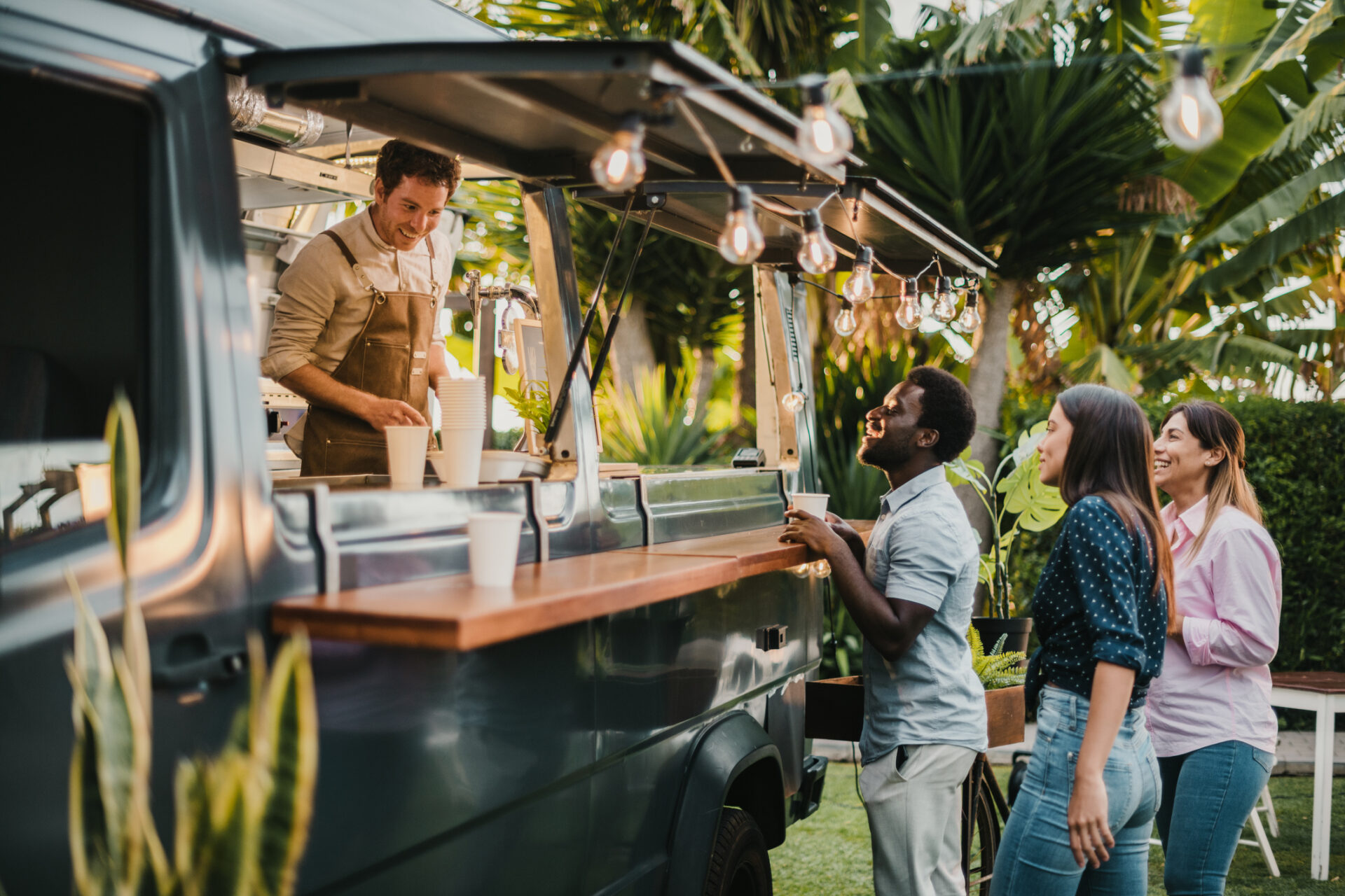 Friends standing in queue and ordering takeaway dishes from man in apron working in food truck.